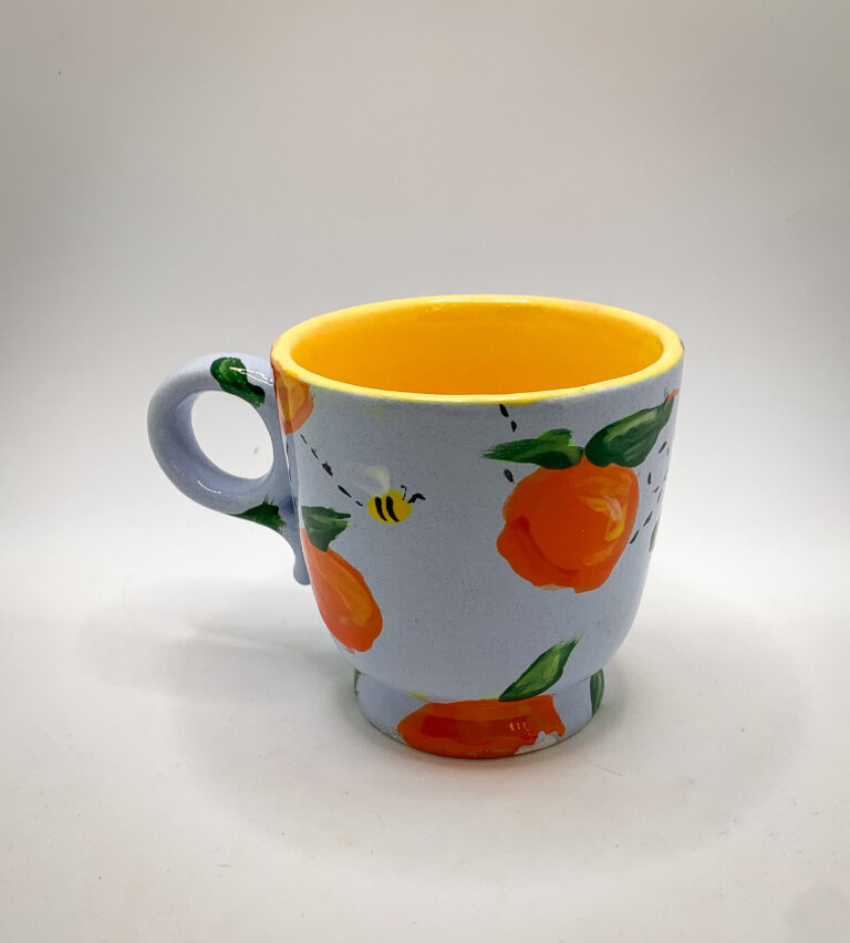 A ceramic mug with a blue exterior featuring hand-painted orange fruit and green leaves. A small bee is also painted near the fruit. The mug's rim and interior are bright yellow.