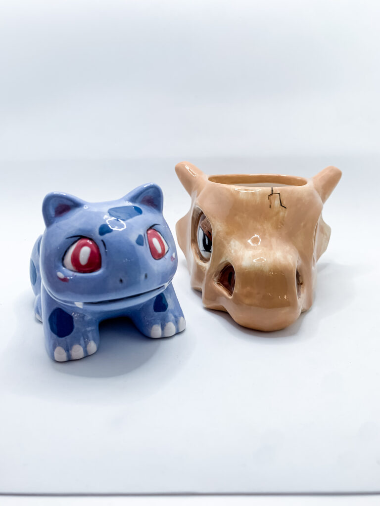 Two ceramic planters shaped like creatures with large eyes. One is blue with spots, resembling a reptile, while the other is light brown with a cracked design on the head, resembling a creature with prominent eye sockets. Both are set against a white background.