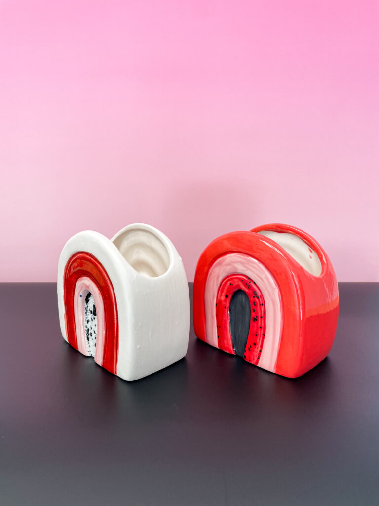 Two ceramic sculptures with arch motifs on a black surface against a pink gradient background. One sculpture is mostly white with red and black details, and the other is predominantly red with pink and black accents.