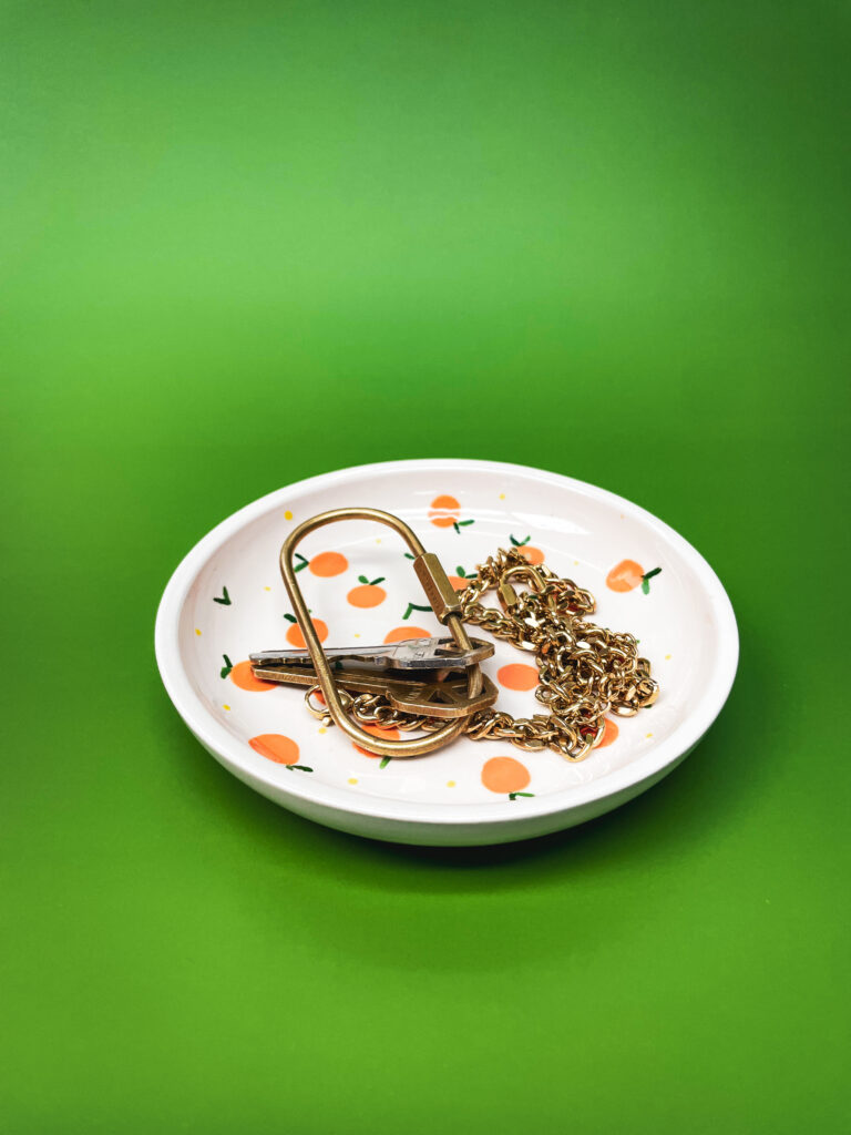 A white dish with orange fruit patterns holds a gold keychain, a set of keys, and a gold chain. The background is a vivid green.