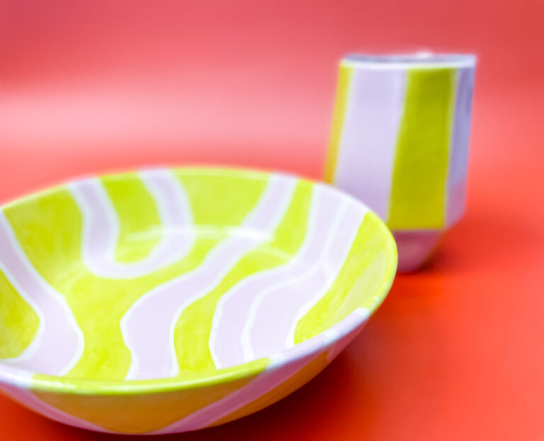 A ceramic bowl and a matching tumbler with white and yellow wavy stripes sit against a vibrant red background. The bowl and tumbler share a modern, abstract design.
