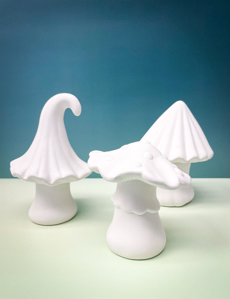 Three white ceramic mushrooms of varying shapes and sizes are displayed on a light green surface, set against a gradient teal background. Each mushroom has a unique cap design, adding a whimsical touch to the arrangement.
