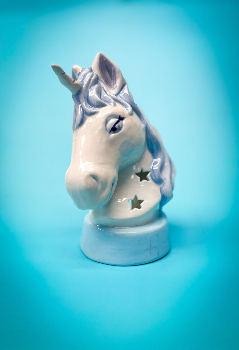 Ceramic unicorn head with a shiny light blue mane, featuring star-shaped cutouts, set against a bright blue background.