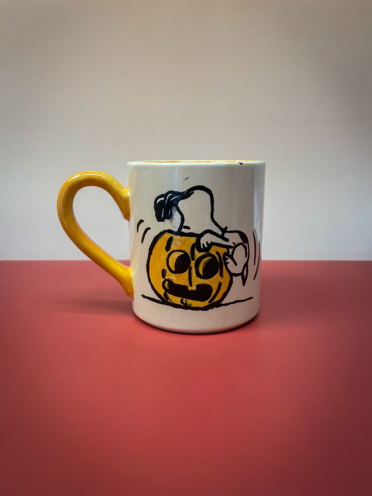 A ceramic mug with a yellow handle sits on a red surface. It features a hand drawing of a ghost with a yellow pumpkin head and comical eyes, surrounded by motion lines. The background is a soft gradient of light to dark.