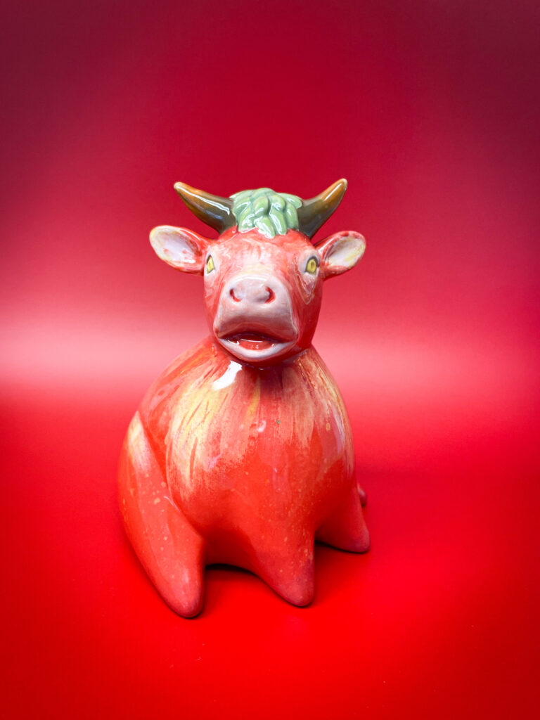 A small, red ceramic cow with large eyes and short horns sitting against a vibrant red background. The cow has a slight grin and a glossy finish, giving it a whimsical and playful appearance.