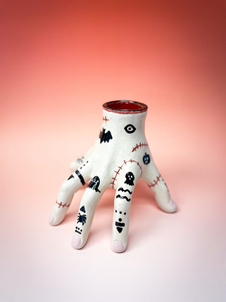 A ceramic vase shaped like a hand with fingers spread out, featuring black and red whimsical designs and patterns on each finger. The background is a gradient of orange to light pink.