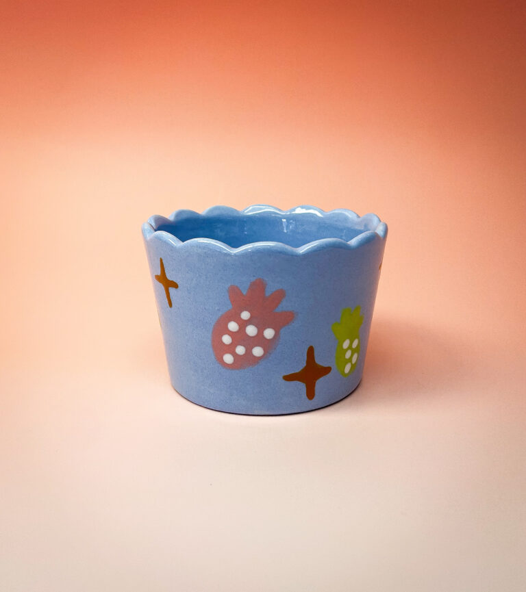 A blue ceramic cup with a scalloped edge is decorated with colorful pineapples and star shapes. The background is a gradient of warm orange and peach tones.
