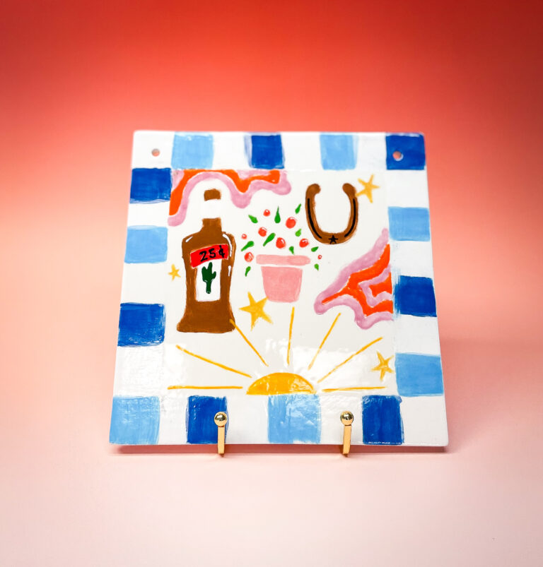 A colorful ceramic tile with blue and white checkered edges features a painted design of a cactus bottle labeled "25A," a glass with stars, a horseshoe, and a sunrise. Two hooks protrude from the bottom, set against a gradient orange background.