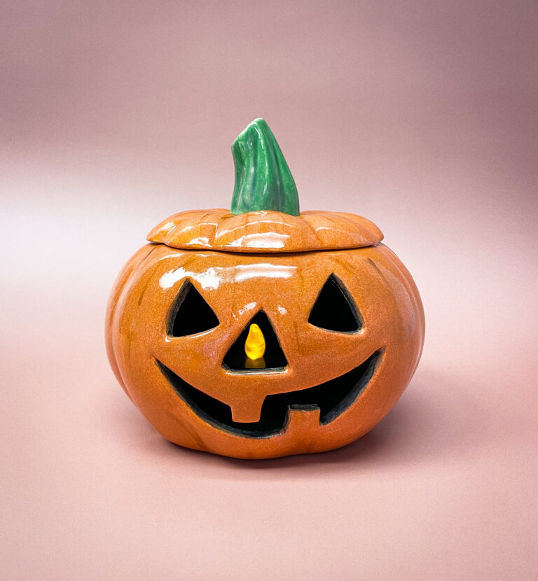 A ceramic jack-o'-lantern with a smiling face, featuring a green stem and a warm, glowing light inside, set against a soft pink background.