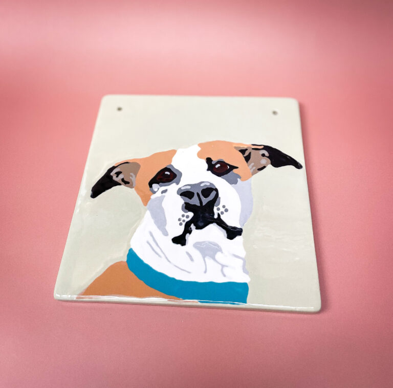 A painted ceramic tile featuring a portrait of a brown and white dog with black ears and a blue collar. The dog is depicted on a light background, and the tile is placed on a pink surface.