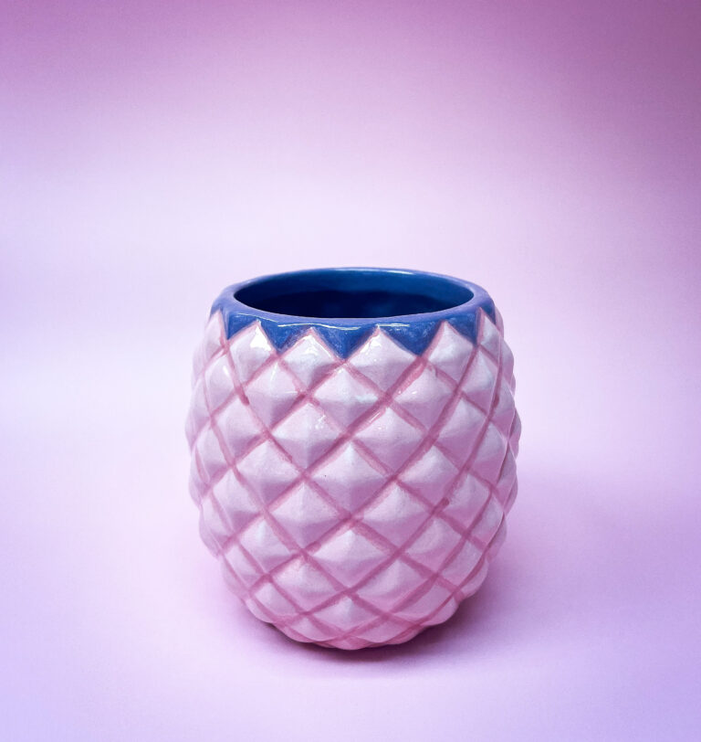A small, textured ceramic pot with a quilted pattern, pink exterior, and blue interior set against a soft pink background.