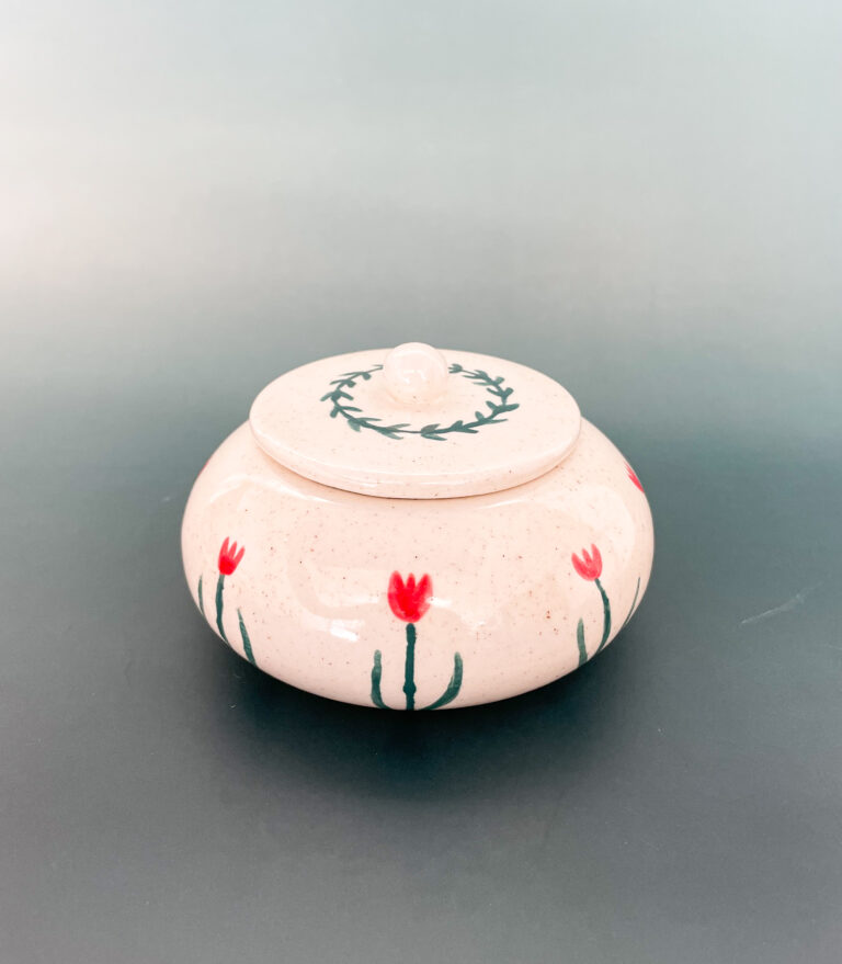 A round, cream-colored ceramic jar with a lid, adorned with hand-painted red flowers and green stems. The lid features a circular green leaf pattern. The jar is set against a soft green gradient background.