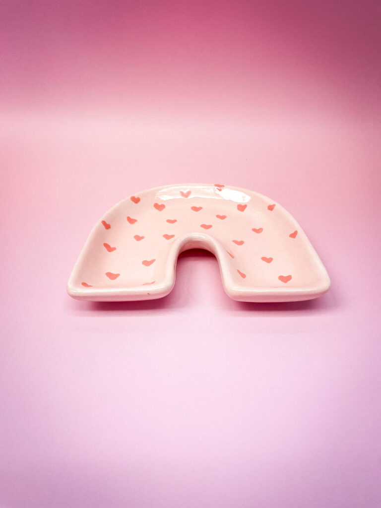 A pink, arch-shaped ceramic dish with small red heart patterns is placed on a gradient pink background.