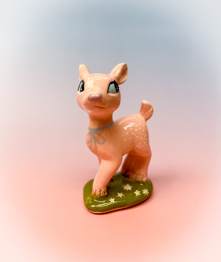 A small ceramic deer figurine stands on a green base adorned with white and yellow flowers. The deer is light brown with white spots and has large, expressive eyes. The background is a soft gradient of pink and blue.