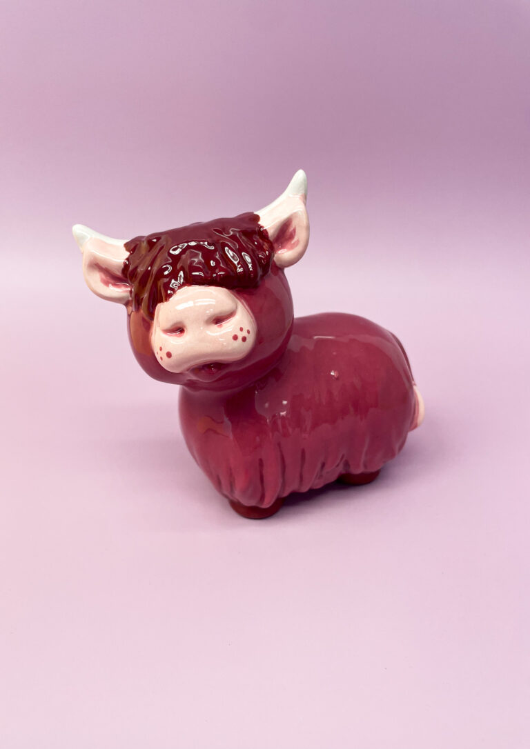 A ceramic figurine of a purple highland cow with white horns and a pink snout is set against a light purple background. The cow has a rounded body and textured fur, giving it a whimsical and playful appearance.