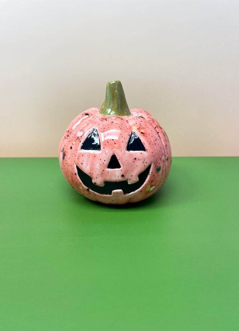 A small, pink ceramic pumpkin with a carved Jack-o'-lantern face sits on a green surface against a beige background. Its stem is painted green.