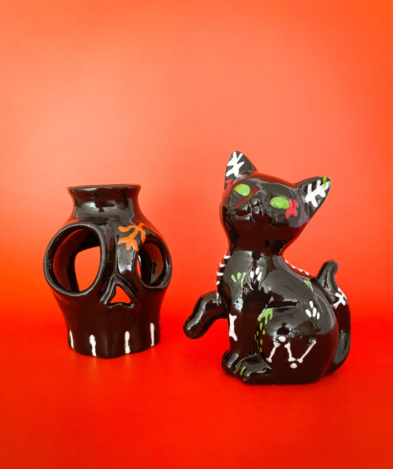 A black decorative skull vase with orange accents and a black ceramic cat with colorful floral patterns are set against a bright orange background.