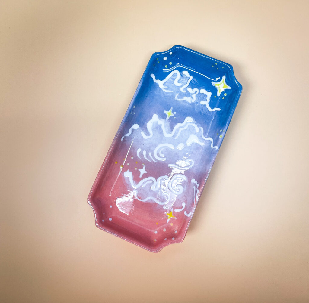 A rectangular tray with rounded corners featuring a gradient from pink to blue. It is decorated with white abstract designs and yellow stars against a beige background.