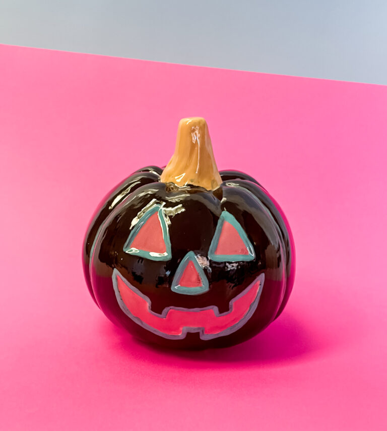A glossy ceramic pumpkin with a carved face is set against a pink background. It features a brown stem, and its eyes, nose, and mouth are highlighted in turquoise.
