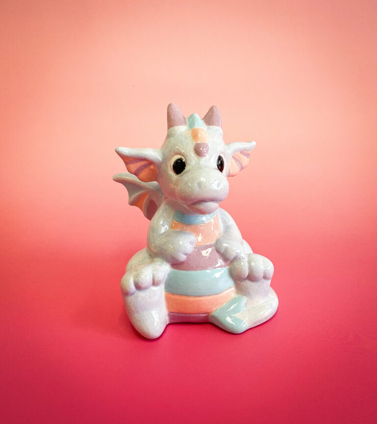 A shiny ceramic dragon figurine with pastel colors sits on a pink gradient background. The dragon has large eyes, small wings, and a striped body, with colors including light pink, blue, and beige.