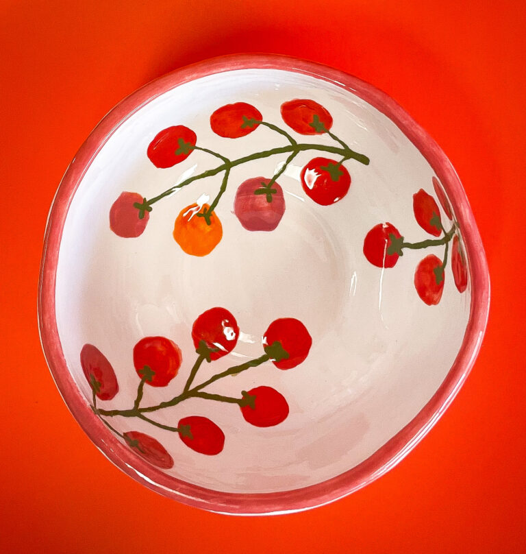 A ceramic bowl with a white interior featuring painted red cherry tomato vines. The rim is red, complementing the vibrant orange background.