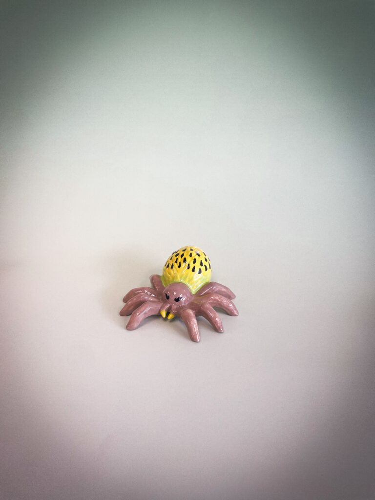 A small, colorful ceramic spider on a smooth, light-colored background. The spider has a yellow, spotted body and pink legs, with a slightly shiny surface, giving it a playful appearance.