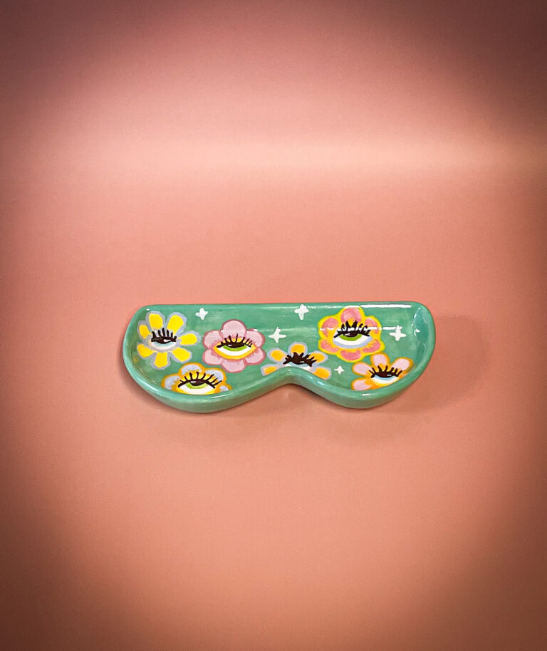 A ceramic mask with a green background, adorned with colorful flower designs and painted eyelashes, is displayed against a pink background. The mask has a whimsical and artistic appearance.