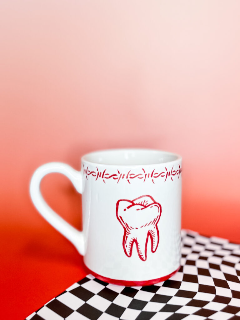 A white mug with a red tooth illustration and barbed wire design sits on a black and white checkered surface. The background is a gradient of pink to red.
