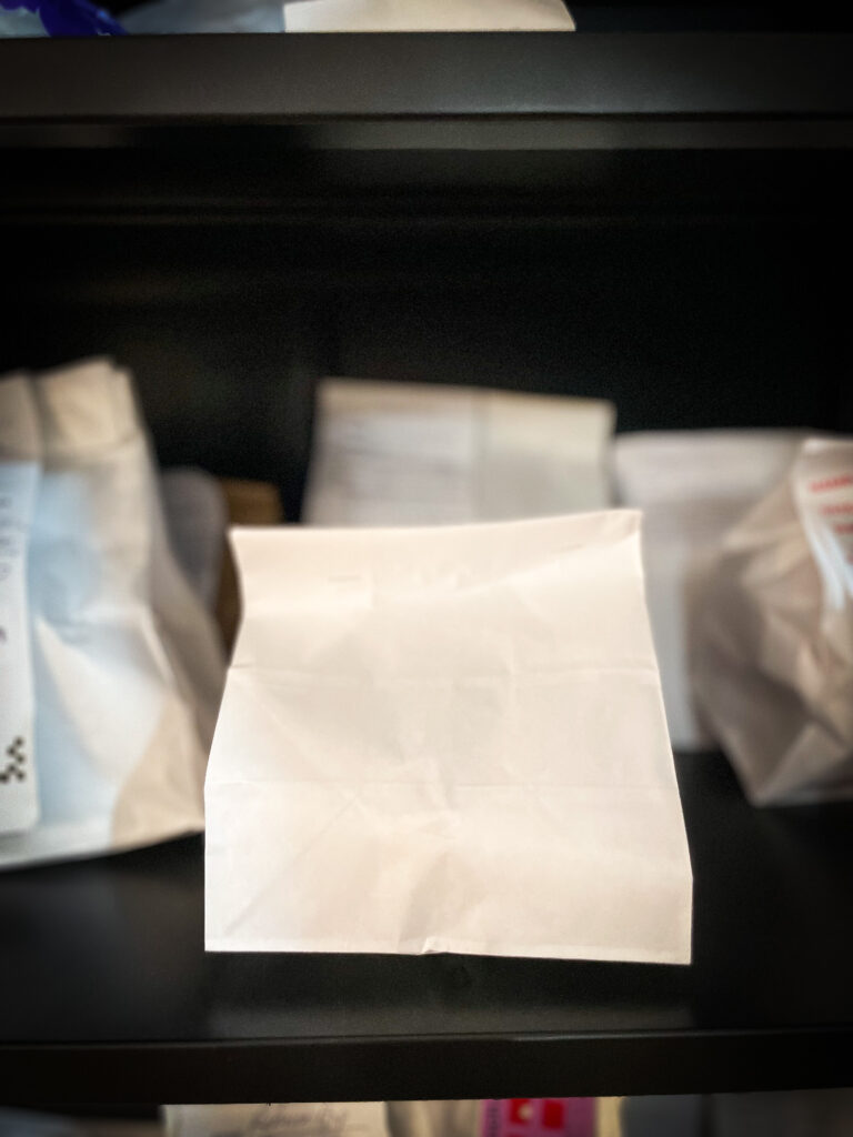 A close-up of several white paper bags on a dark shelf. The bags are unmarked and appear to be neatly arranged in a row, with one bag prominently in the foreground.