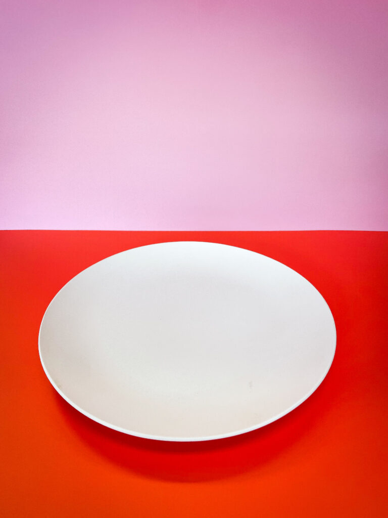 A white, round plate sits empty on a vibrant red surface. The background fades from pink to light pink, creating a colorful contrast with the plate.
