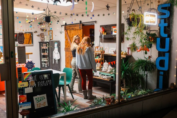 Two people stand inside a cozy pottery studio in the Twin Ports area, decorated with hanging ornaments and plants. A sign outside shows operating hours as 12-8. Various pottery items are displayed on a shelf. Bright colors and lights create a welcoming atmosphere, making it one of the fun things to do in Duluth, MN.