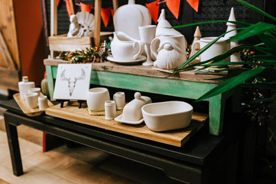 A collection of unpainted ceramic items, including mugs, bowls, and figurines from Brush & Co., is displayed on a green and wooden shelf. A small easel holds a card with a reindeer antler design. Green leaves partially frame the scene, and red flags hang above—a charming spot for fun things to do in Superior, Wisconsin.