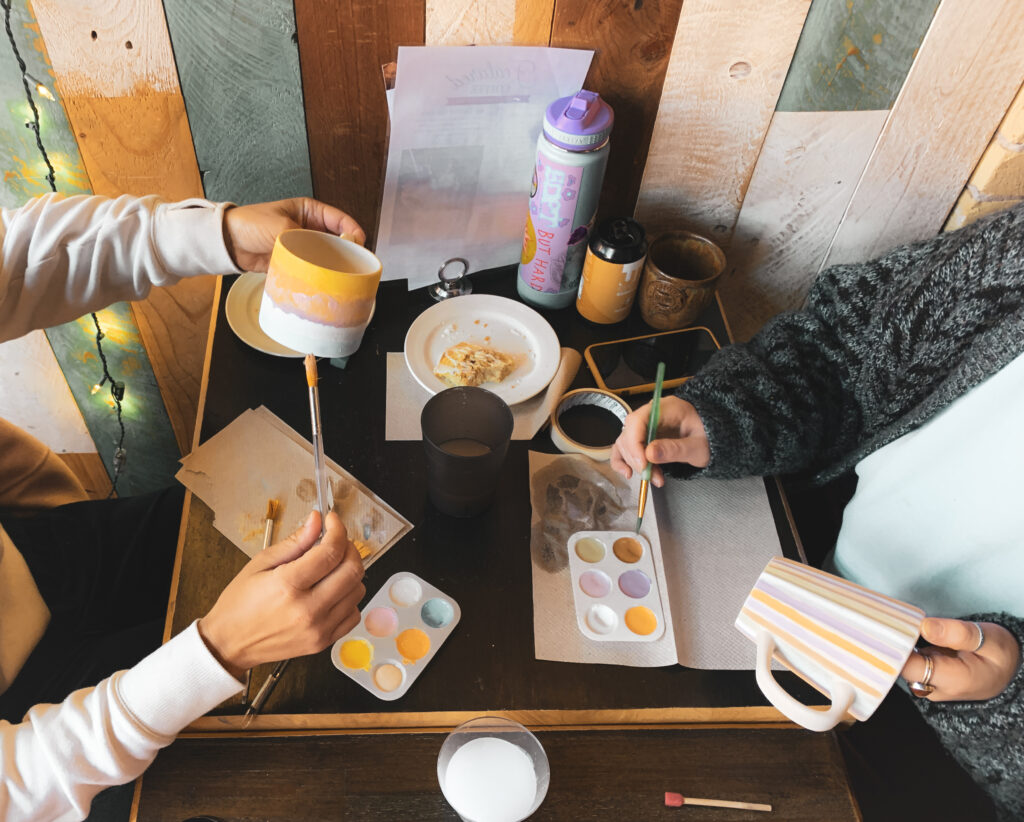 Two people sit at a table painting pottery mugs with pastel colors, surrounded by a delightful mess of Brush & Co.'s paint palettes, brushes, cups, and some plates. The background features multi-colored wooden paneling, adding to the creative ambiance.