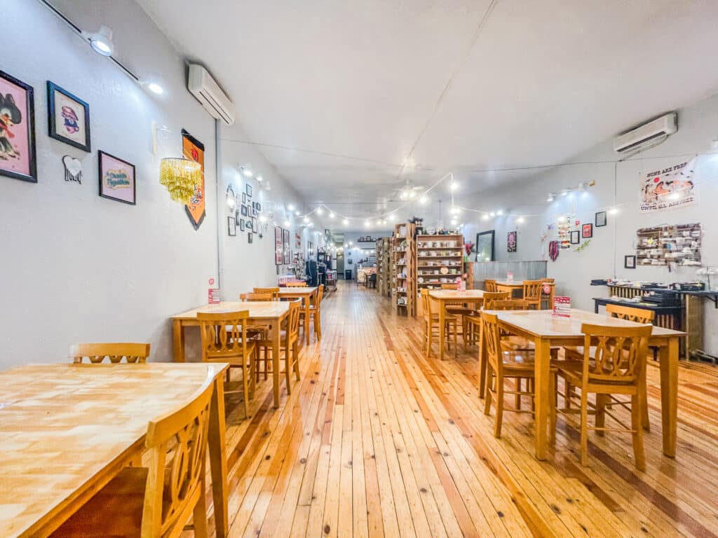 Spacious, brightly lit café by Brush and Co. with wooden floors and tables, wooden chairs, string lights overhead, and artwork decorating the walls; shelves and decorations visible in the background.