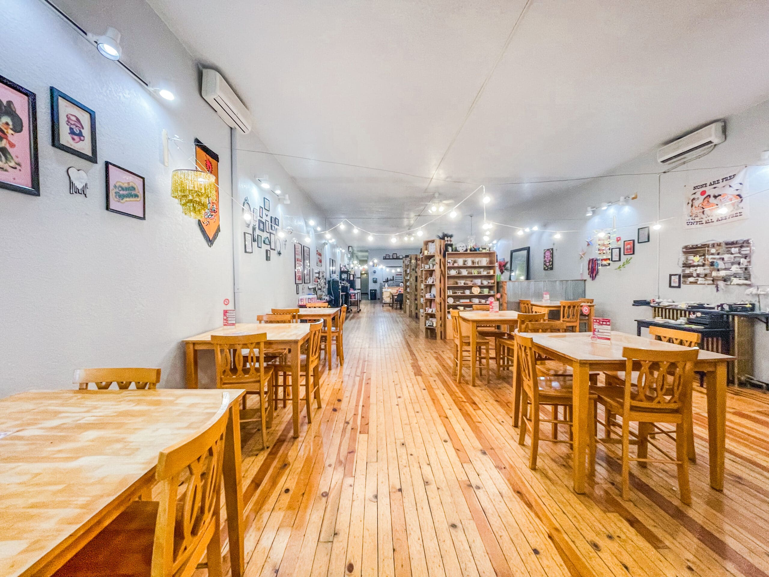 Spacious, brightly lit café by Brush and Co. with wooden floors and tables, wooden chairs, string lights overhead, and artwork decorating the walls; shelves and decorations visible in the background.