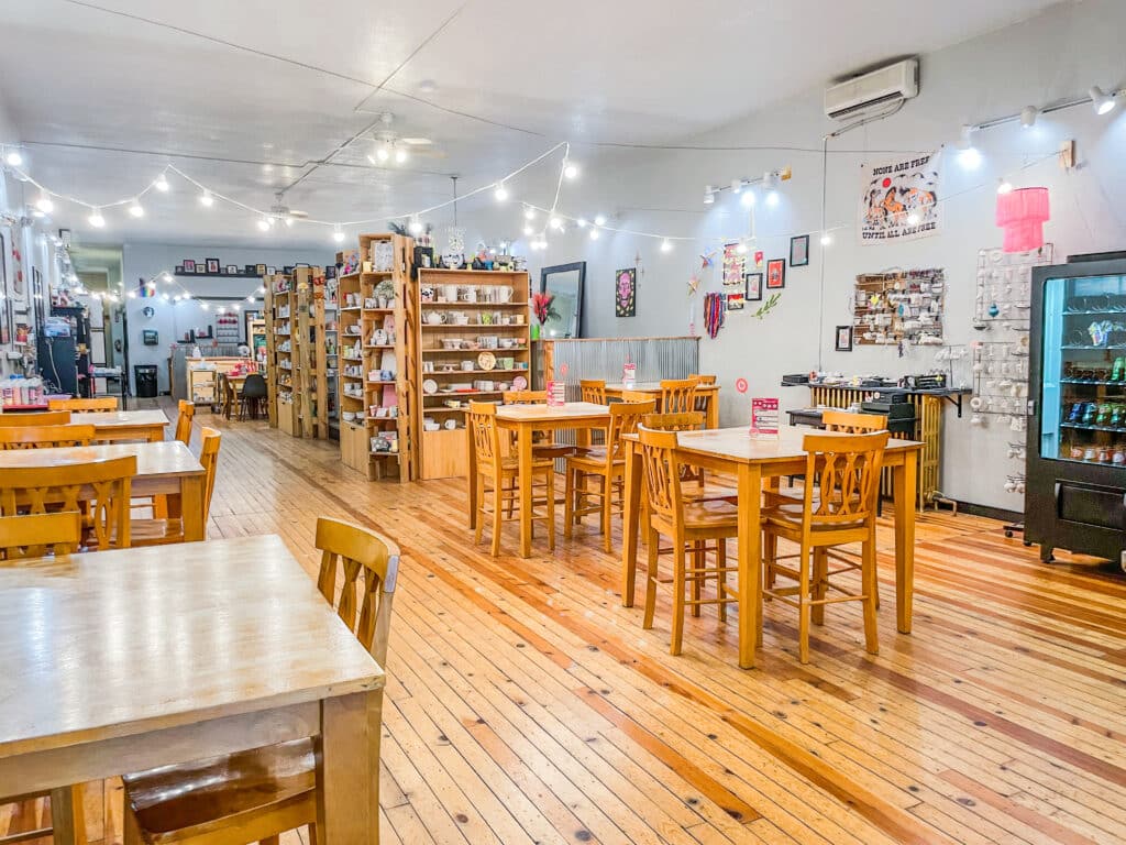 A cozy café by Brush and Co., with wooden tables and chairs, shelves filled with mugs and décor, fairy lights hanging from the ceiling, and various artwork and posters on the white walls. The atmosphere is warm and inviting.