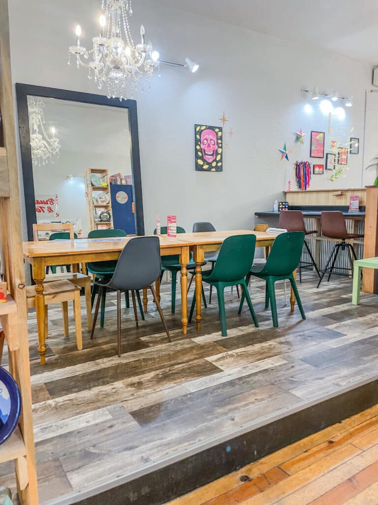 A cozy, brightly lit Brush & Co. art studio with a large wooden table surrounded by mismatched chairs, colorful wall art, a large mirror, and a chandelier hanging from the ceiling. Art supplies and decorations are visible throughout the room.