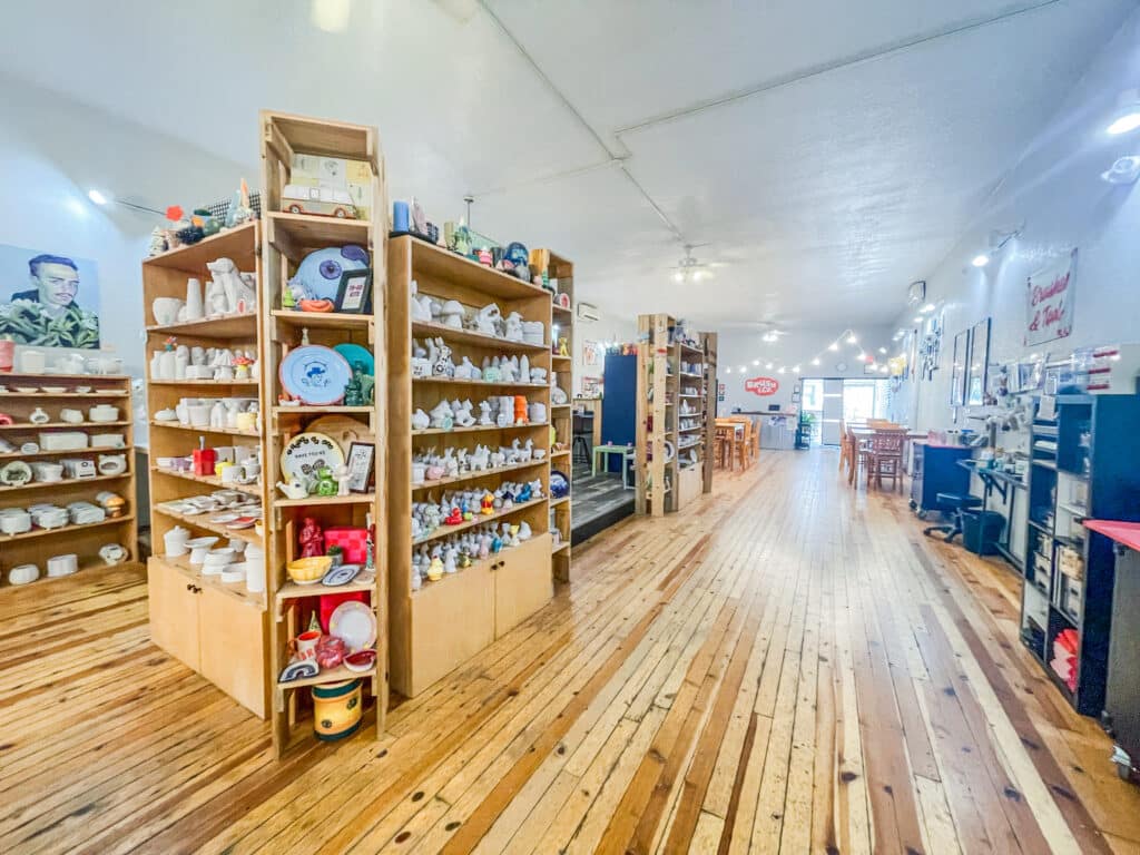 A bright, spacious studio by Brush & Co. features wooden floors and shelves filled with pottery and ceramic items. Tables and chairs are set up for painting, with string lights overhead creating a welcoming atmosphere.