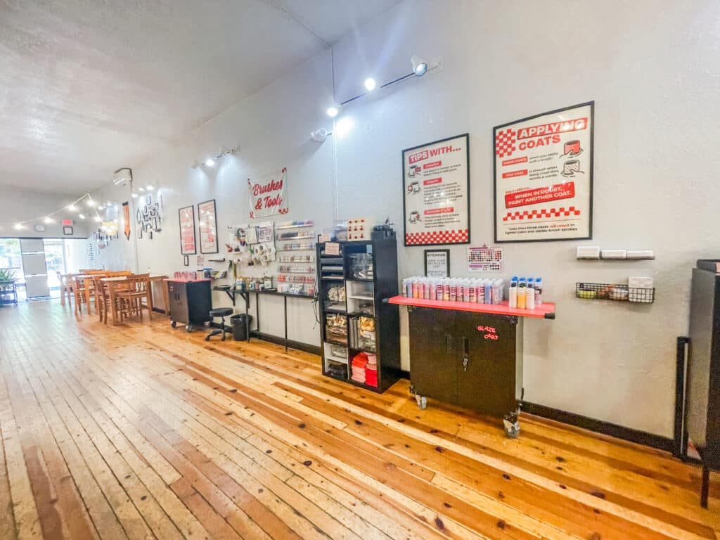 A chic Brush & Co. nail salon interior with wooden floors, a long wall of nail products, instruction posters, a desk and chair, plus a row of tables and chairs bathed in natural light near the bright window at the far end.